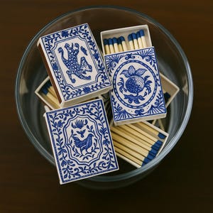 May include: A collection of decorative matchboxes with blue and white designs, including a leopard, a fruit, and a bird. The matchboxes are inside a clear glass bowl, with the match heads painted blue.