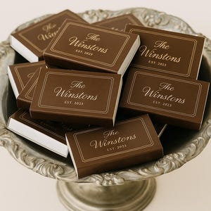 Può includere: Una collezione di scatole rettangolari marroni con la scritta dorata "The Winstons EST. 2023". Le scatole sono disposte su un vassoio d'argento decorativo.