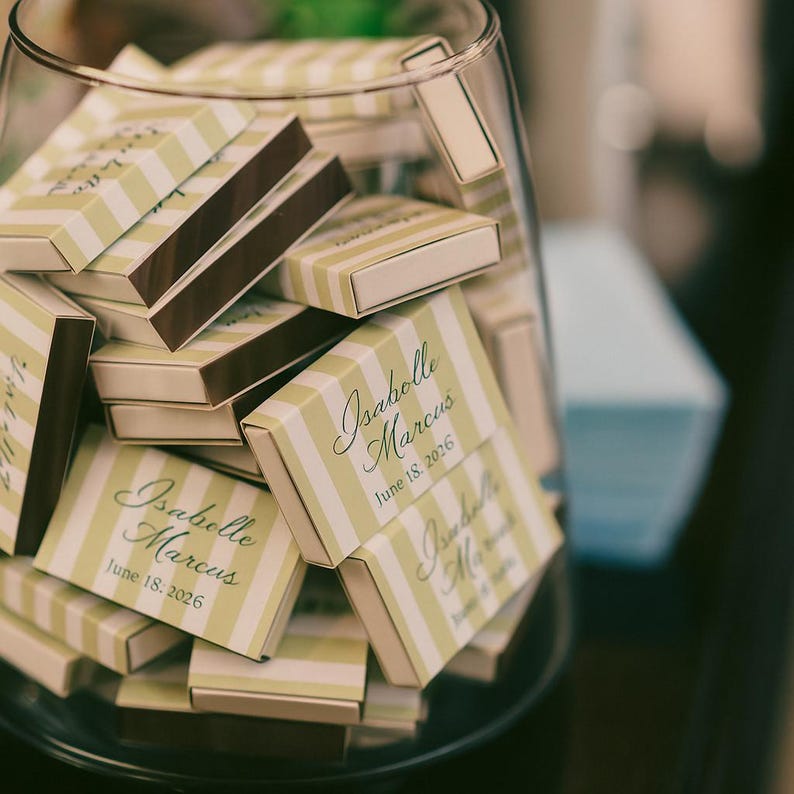 May include: A clear glass jar filled with small matchboxes. The matchboxes have a light green and white striped pattern with a dark brown striking surface. The text "Isabelle Marcus June 18, 2026" is printed on the matchboxes.