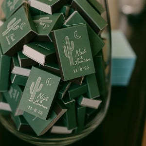 May include: A glass jar filled with green matchboxes. Each matchbox features a white cactus and crescent moon design, along with the names "Nick & Lauren" and the date "11-8-25". The matchboxes are a wedding favor.