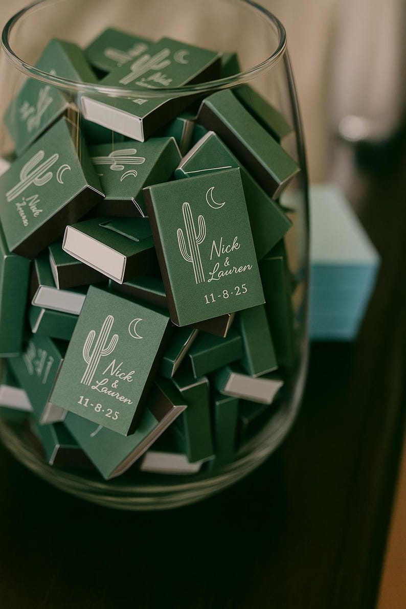 May include: A glass jar filled with green matchboxes. Each matchbox features a white cactus and crescent moon design, along with the names "Nick & Lauren" and the date "11-8-25". The matchboxes are a wedding favor.