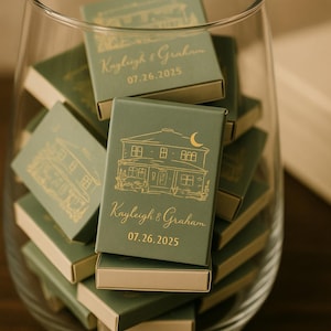May include: A collection of green matchboxes with gold foil details inside a clear glass container. Each box features a house illustration and the names "Kayleigh & Graham" with the date "07.26.2025".