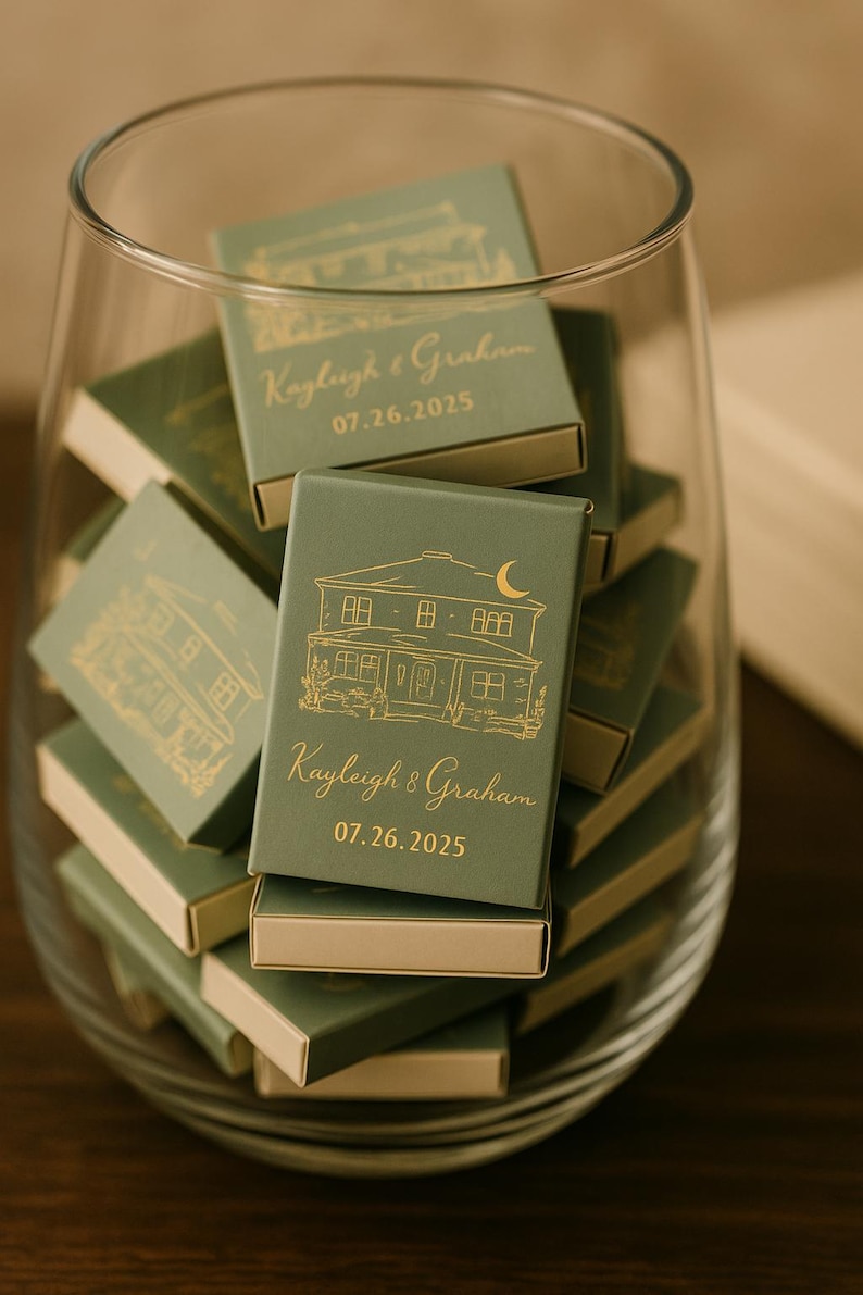May include: A collection of green matchboxes with gold foil details inside a clear glass container. Each box features a house illustration and the names "Kayleigh & Graham" with the date "07.26.2025".