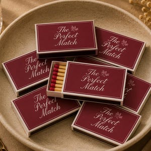 Pode incluir: Caixas de fósforos bordô com a inscrição "The Perfect Match" em escrita branca, dispostas sobre um prato bege. Uma caixa está aberta, mostrando os fósforos. Uma vela acesa e uma corrente dourada são visíveis no fundo.