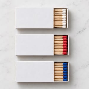 May include: Three rectangular matchboxes with plain white sliding covers. Each box contains wooden matches with distinct colored tips: the top box has white tips, the middle box has red tips, and the bottom box has blue tips, all arranged in rows.