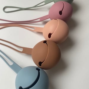 May include: A collection of five silicone pacifier holders in various pastel colors: green, pink, peach, brown, and blue. Each holder has a round shape with a slit and a loop for attaching to a pacifier or other item.