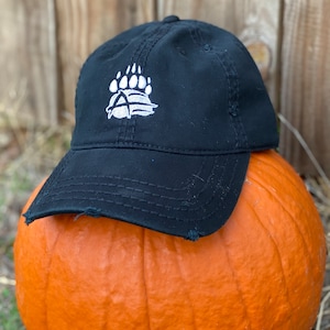 Pode incluir: Um boné de beisebol preto desgastado com um logotipo de pata de urso bordado em branco. O boné está em cima de uma abóbora laranja.