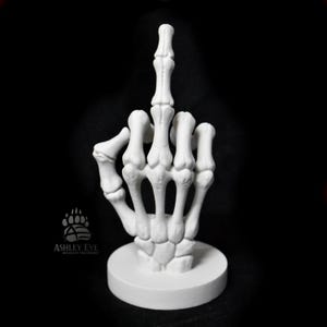 May include: A white, skeletal hand sculpture giving the middle finger. The hand is mounted on a circular base. The sculpture is set against a black background. The sculpture is a novelty item.