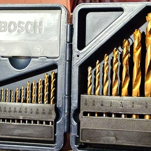 May include: An open, blue plastic case containing a set of gold-colored drill bits. The case is labeled "BOSCH" on the lid. The drill bits are arranged in size order, with measurements visible on the holders. A practical tool for various drilling tasks.