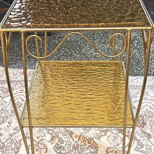 May include: A vintage gold-colored side table with two textured glass shelves. The table has a decorative, curved design on the front and is supported by slender, gold-toned metal legs. The table's dimensions are approximately 24 inches high, 18 inches wide, and 12 inches deep.