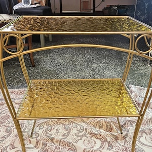 May include: A gold-colored, two-tiered side table with a textured glass top and shelf. The table has a curved metal frame with decorative circular accents. The table's legs are thin and tapered.