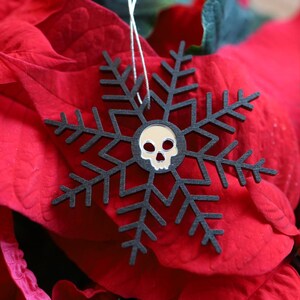 Può includere: Un ornamento a forma di fiocco di neve nero con un design di teschio bianco al centro, appeso a una pianta di stella di Natale rossa.