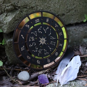 K&ouml;nnte beinhalten: Ein h&ouml;lzernes Zodiakrad mit schwarzem Hintergrund und goldenen Akzenten. Das Rad zeigt die zw&ouml;lf Tierkreiszeichen und Sternbilder. Das Rad ist von einem goldenen Ring umgeben.
