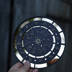 K&ouml;nnte beinhalten: Ein schwarzes, rundes Holzschild mit einer verspiegelten Oberfl&auml;che und einem goldenen Tierkreisrad mit Sternbildern und Sternzeichen. Das Schild wird in einer Hand gehalten.