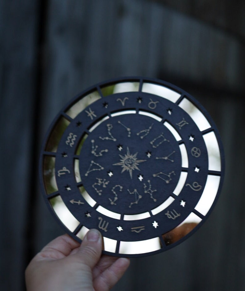 K&ouml;nnte beinhalten: Ein schwarzes, rundes Holzschild mit einer verspiegelten Oberfl&auml;che und einem goldenen Tierkreisrad mit Sternbildern und Sternzeichen. Das Schild wird in einer Hand gehalten.