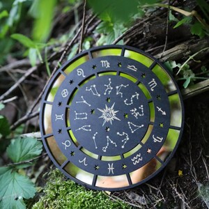 K&ouml;nnte beinhalten: Ein schwarzes Holz-Zodiakrad mit goldenen Akzenten und den Sternbildern der Tierkreiszeichen. Das Rad ist von einem goldenen Hintergrund umgeben und liegt auf einem Bett aus Moos und gr&uuml;nen Bl&auml;ttern.