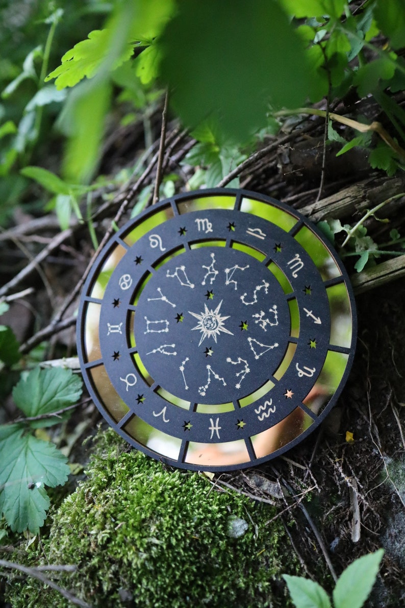K&ouml;nnte beinhalten: Ein schwarzes Holz-Zodiakrad mit goldenen Akzenten und den Sternbildern der Tierkreiszeichen. Das Rad ist von einem goldenen Hintergrund umgeben und liegt auf einem Bett aus Moos und gr&uuml;nen Bl&auml;ttern.