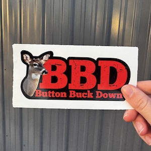 May include: A white sticker with the text "BBD Button Buck Down" in red letters with a black outline. A deer head graphic is on the left side of the sticker. The sticker is held by a hand.