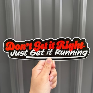 May include: A black and white sticker with the text "Don't Get it Right - Just Get it Running" in red and white lettering. The sticker is held up by a hand against a gray background.