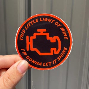 May include: A round sticker with a black background and an orange engine symbol. The sticker has the text "THIS LITTLE LIGHT OF MINE I'M GONNA LET IT SHINE" in orange around the edge. The sticker is held up by a hand.