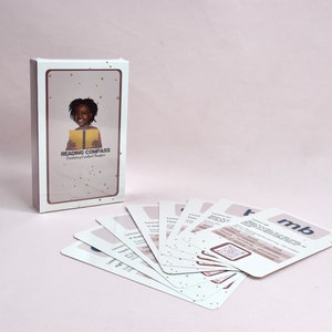 May include: A box of Reading Compass Developing Confident Readers cards with a colorful illustration of a young girl reading a book. The cards are spread out on a pink surface.