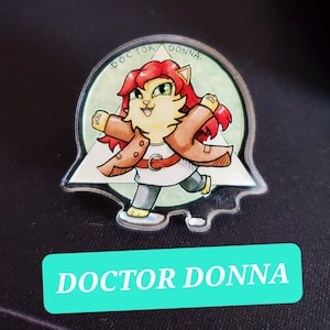May include: A clear acrylic pin featuring a cartoon illustration of a red-haired cat wearing a brown coat and a white shirt. The cat is walking and has a green background. The text "DOCTOR DONNA" is written around the edge of the pin.