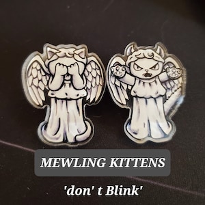 Op de afbeelding: Twee witte engelbeeldjes met kattenkoppen. De engel links bedekt zijn ogen met zijn handen. De engel rechts heeft een duivelse uitdrukking en houdt zijn handen dreigend omhoog. De tekst "MEWLING KITTENS 'don't Blink'" staat onder de beeldjes.