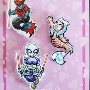 May include: Three colorful mermaid character pins with red, blue, and purple tails. The characters are holding lightsabers. The pins are on a pink background with white paw prints. The text "www.NicDaStick.com" is at the bottom of the image.