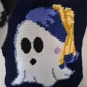 May include: Crocheted blanket featuring a cartoon ghost with black eyes and mouth, blue hair, and yellow accents. The ghost is set against a navy blue background. The blanket is handmade with visible stitches.