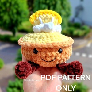 Peut inclure: Une peluche en crochet en forme de tarte avec un visage souriant, une garniture jaune et une garniture blanche en forme de nuage. La tarte a une croûte brune et des pieds bordeaux. Le texte "PDF PATTERN ONLY" est visible.
