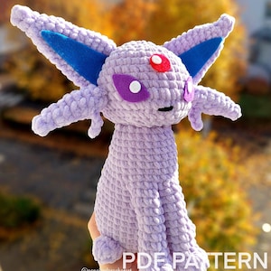 Peut inclure: Peluche Espeon au crochet lavande avec des accents d'oreilles bleues, une gemme rouge et des yeux violets. La peluche est assise, les jambes tendues. Le texte "PDF PATTERN ONLY" est en bas.