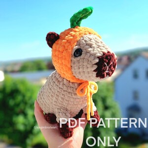 Peut inclure: Une peluche capybara crochetée portant un chapeau citrouille orange avec une tige verte. La peluche est beige avec des accents marron foncé. Le texte "PDF PATTERN ONLY" est visible.
