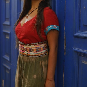 Puede incluir: Una persona con una blusa roja con cuello en V, un cinturón estampado colorido y pantalones verde oliva. Las mangas de la blusa tienen adornos azules y florales. Está apoyada contra una puerta azul.