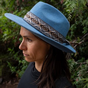 Peut inclure: Un chapeau fedora bleu clair avec une bande décorative à motifs géométriques marron, blancs et noirs. Le chapeau a un large bord et un détail de pompon. Un accessoire élégant.
