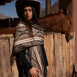 Puede incluir: Una mujer con un poncho marrón y negro con flecos, un sombrero marrón y pantalones negros se encuentra junto a un caballo marrón. La mujer mira hacia la izquierda del encuadre. El caballo está apoyando la cabeza en una valla de madera.