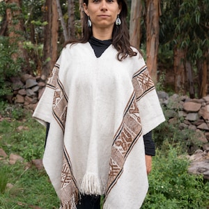 Op de afbeelding: Crèmekleurige poncho met bruine en zwarte geometrische patronen op de mouwen en randen. De poncho heeft een V-hals en franjes langs de onderkant. De persoon draagt een zwarte top en een blauwe jeans.