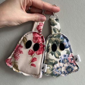 May include: Two ghost-shaped fabric keychains with metal lobster clasps. One keychain features a beige background with pink floral patterns, while the other has a cream background with blue, pink, and green floral prints. Both have black felt eyes.