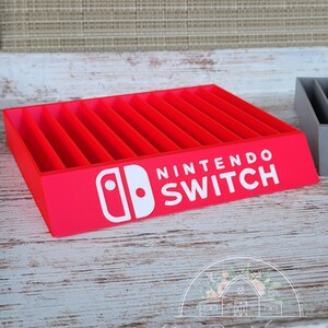 May include: A red Nintendo Switch game cartridge organizer with multiple slots. The white Nintendo Switch logo is prominently displayed on the front. The organizer is made of wood and sits on a white and gray wooden surface.