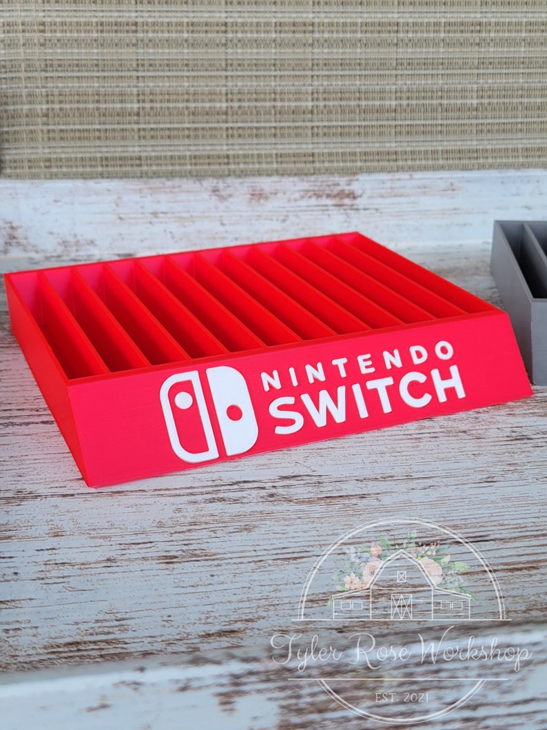May include: A red Nintendo Switch game cartridge organizer with multiple slots. The white Nintendo Switch logo is prominently displayed on the front. The organizer is made of wood and sits on a white and gray wooden surface.