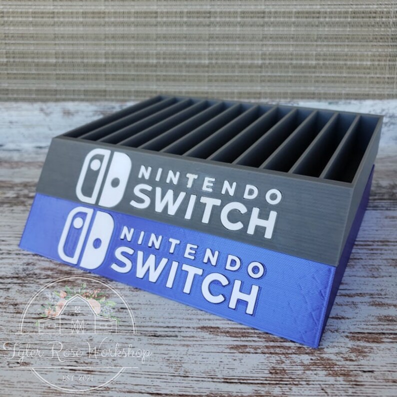 May include: A gray and blue Nintendo Switch game cartridge holder. The holder has multiple slots and features the Nintendo Switch logo and text in white. The base is blue, and the top is gray.