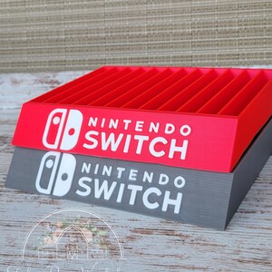 May include: A red and gray Nintendo Switch game holder with the words "NINTENDO SWITCH" in white. The holder has multiple slots for game cartridges. The logo is also visible. The holder is made by Tyler Rose Workshop.