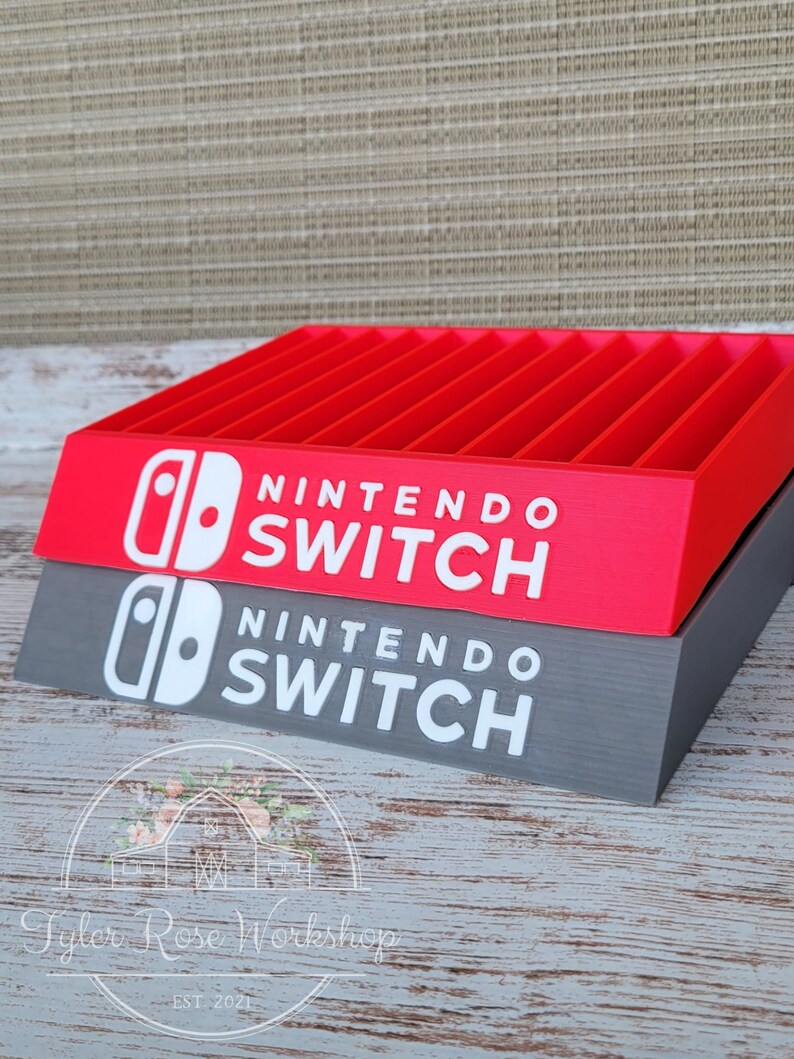 May include: A red and gray Nintendo Switch game holder with the words "NINTENDO SWITCH" in white. The holder has multiple slots for game cartridges. The logo is also visible. The holder is made by Tyler Rose Workshop.