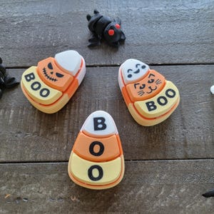 Puede incluir: Tres adornos de maíz dulce con temática de Halloween con la palabra "BOO" en cada uno. Los adornos son blancos, naranjas y amarillos.