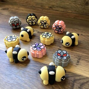 May include: A set of 10 bee-themed 3D printed cookie cutters. The cutters are in various colors, including yellow, pink, gray, and black. The cutters feature different bee designs, including a bee with wings, a bee with a honeycomb, and a bee with a flower.