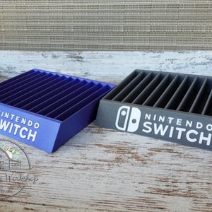 May include: Two Nintendo Switch game organizers, one blue and one gray, each with slots for game cartridges. The Nintendo Switch logo and text are printed in white on the front of each organizer. The organizers are displayed on a wooden surface.