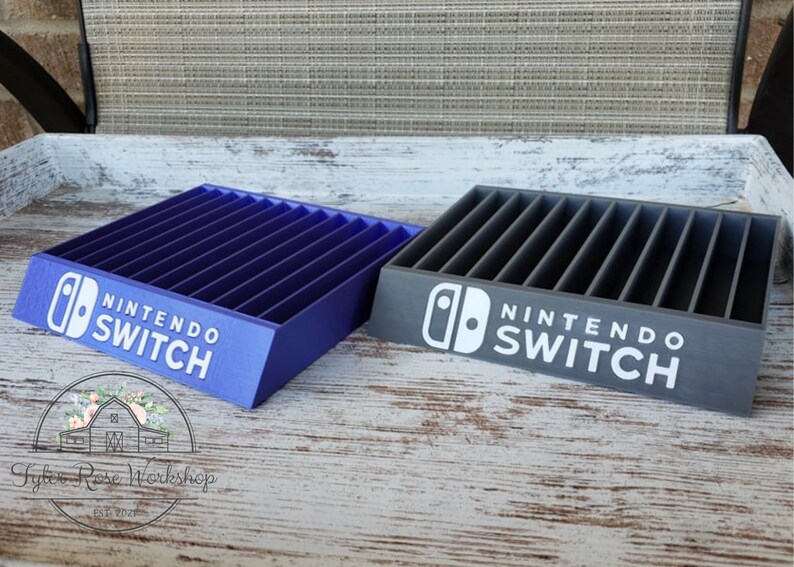 May include: Two Nintendo Switch game organizers, one blue and one gray, each with slots for game cartridges. The Nintendo Switch logo and text are printed in white on the front of each organizer. The organizers are displayed on a wooden surface.