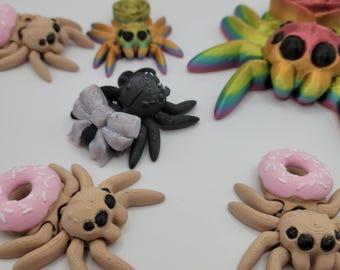 May include: A collection of 3D printed spider figurines with various colors and designs. The spiders are made of plastic and have a variety of details, including eyes, legs, and a bow. The spiders are all different sizes and shapes.