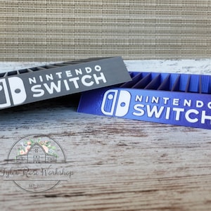 May include: Two Nintendo Switch game organizers, one gray and one purple, are displayed. Each organizer has slots for game cartridges and features the "Nintendo Switch" logo in white. The organizers are on a wooden surface.