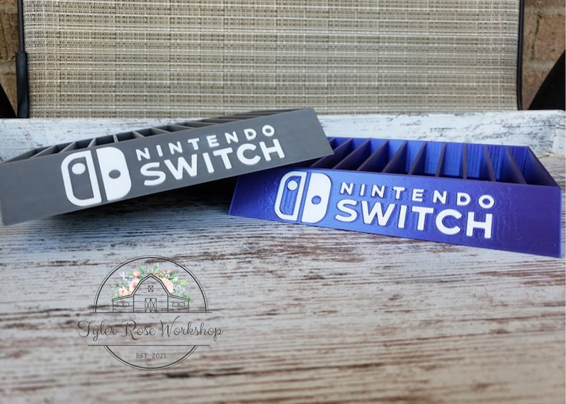 May include: Two Nintendo Switch game organizers, one gray and one purple, are displayed. Each organizer has slots for game cartridges and features the "Nintendo Switch" logo in white. The organizers are on a wooden surface.