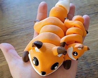 May include: A pair of orange and brown clay fox figurines. The larger fox is curled up with its tail wrapped around its body. The smaller fox is curled up on top of the larger fox. Both foxes have black eyes and white accents.